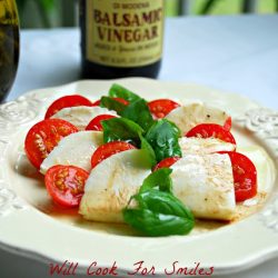 classic caprese salad on a decorative white plate with a bottle of balsamic vinegar and a jar of olive oil in the background while all are on a table with white cloth