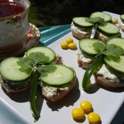 3 cucumber tea shamrock sandwiches on a white plate with a blue table cloth below and a cup of tea in a decorative tea mug to the left.