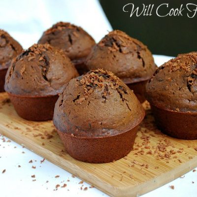 6 chocolate zucchini muffins on a wood cutting board on white table with chocolate shaving around muffins
