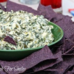 a bed of blue tortilla chips on a platter with a green bowl filled with spinach parmesan dip in the center of chips
