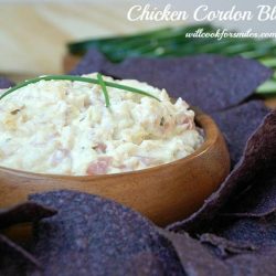 chicken cordon bleu dip in a wooden bowl with blue chips around bowl, cucumbers slice in background and a bottle of red style beer in background to the left