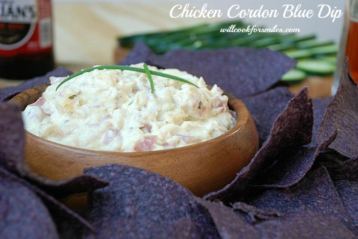 chicken cordon bleu dip in a wooden bowl with blue chips around bowl, cucumbers slice in background and a bottle of red style beer in background to the left