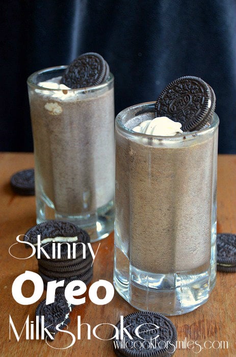 two Oreo Milkshake in a glass mug with whipped cream and an oreo as garnish on a wood table with oreos all around it 