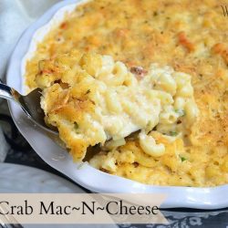 shot of baked crab mac n cheese in white baking dish with spoon in dish left of plate