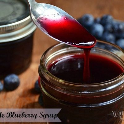 2 mason jars with blueberry syrup one with lid on, 1 spoon in syrup and blueberries on table