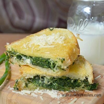 SPinach feta parmesan grilled cheese cut in half staked one each other with 1 greenonion to the left resting on wood cutting board glass of milk in back ground