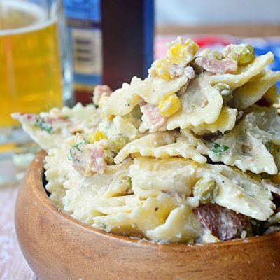 shot from front pasta salad in wood bowl on wood table with glass resting behind in background