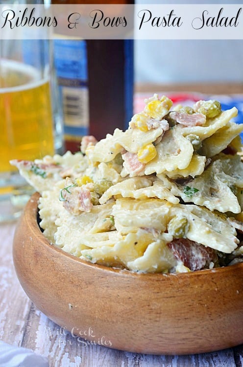 shot from front pasta salad in wood bowl on wood table with glass resting behind in background