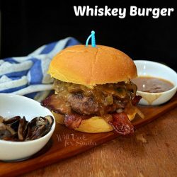 1 whiskey burger in wood cutting board with small white bowl of mushroom to the left, white bowl of sauce to the right and a white and blue towel in background