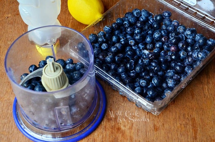 Blueberries in a food processer with the lid off and a plastic bin of blueberries to the right of it 