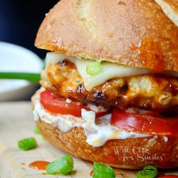buffalo chicken burger on wood cutting board with green onion and tomato in background and small white bowl of ranch