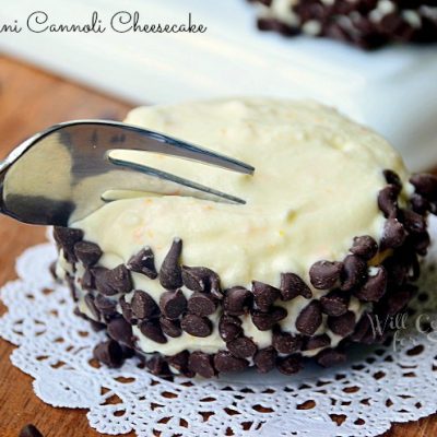 fork cutting into cannoli cheesecake on decorative white napkin with white rectangular plate and additional cannolis in background all on wooden table