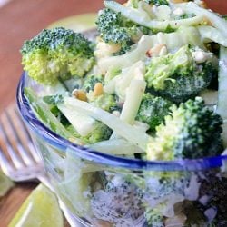 clear glass bowl filled with broccoli cucumber salad on wood cutting board resting on wooden table with fork and limes to the left and right