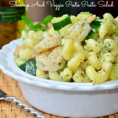 decorative white bowl filed with shrimp and veggie pesto pasta salad on wicker placemat and wooden table with fork to left of the bowl