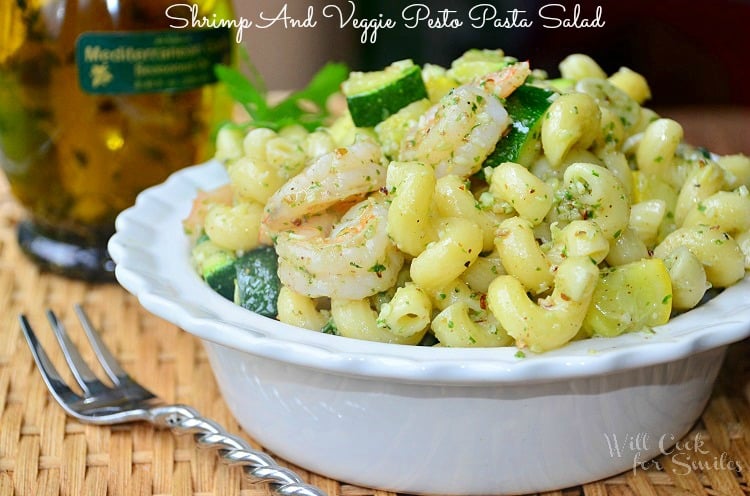 decorative white bowl filed with shrimp and veggie pesto pasta salad on wicker placemat and wooden table with fork to left of the bowl