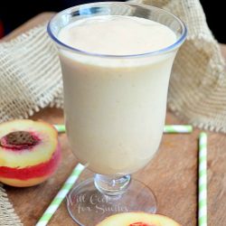 dessert glass filled with skinny peaches and cream milkshake on wooden table with green and white straws and peaches surrounded by burlap cloth