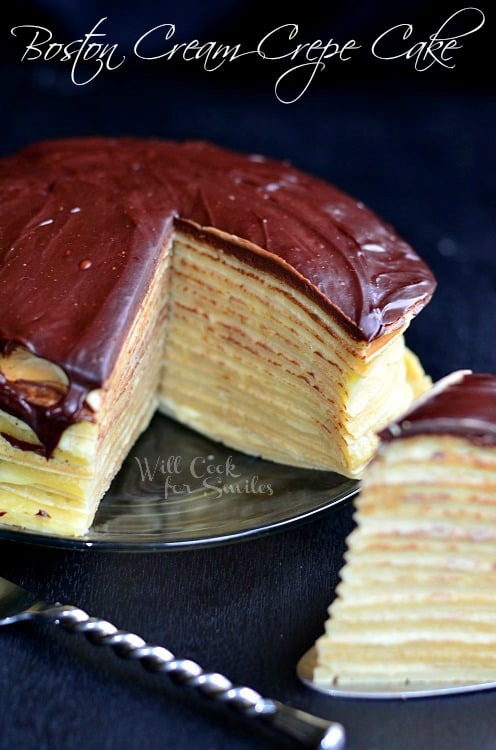 Boston Cream Crepe Cake slice on a cake cutter on a black counter top and the rest of the cake to the top left 