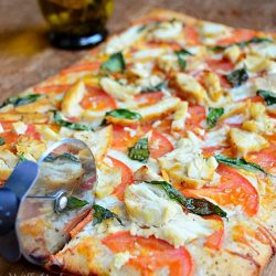 whole caprese chicken pizza on wood cutting board with bottle of olive oil in background to the left with pizza cutter slicing