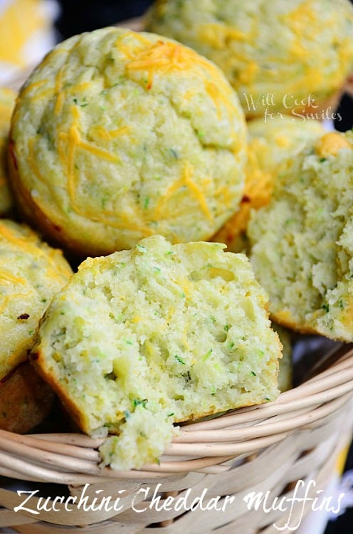 Cheddar-Zucchini Muffins in a wicker type basket. One muffin is torn in half showing the inside of the muffin. Green flecks and cheese are seen throughout.