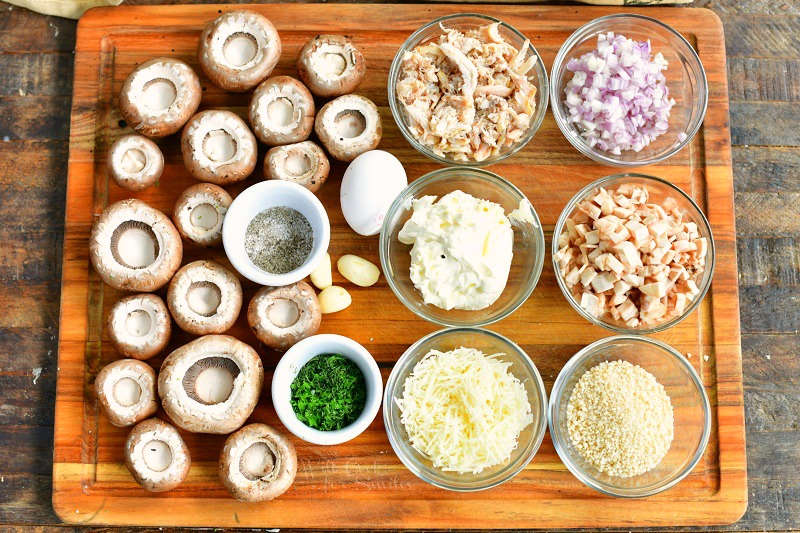 ingredients laid out on the wooden board: mushroom caps, salt and pepper, minced parsley, garlic cloves, crab meat, mayo, parmesan cheese, onion, mushrooms caps, and bread crumbs