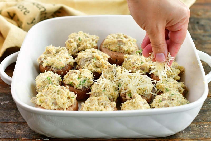 sprinkling shredded Parmesan cheese over stuffed mushrooms uncooked in a white baking dish
