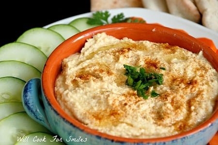 Hummus in a blue and orange bowl with cucumber slices to the left 