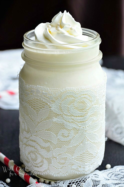 White Wedding Cake Milkshake in a mason jar with lace around the bottom of the mason jar and whipped cream and candy pearls on top with a red and white straws next to it 