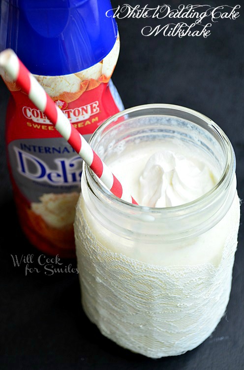 White Wedding Cake Milkshake in a mason jar with lace around the bottom of the mason jar and whipped cream and candy pearls on top with a red and white straw