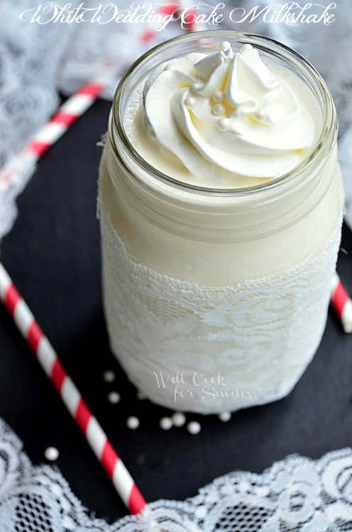 White Wedding Cake Milkshake in a mason jar with lace around the bottom of the mason jar and whipped cream and candy pearls on top with a red and white straws next to it 