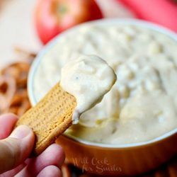 hand holding cookie with dip on it above brown bowl filled with apple pie dip surrounded by cookies and pretzels