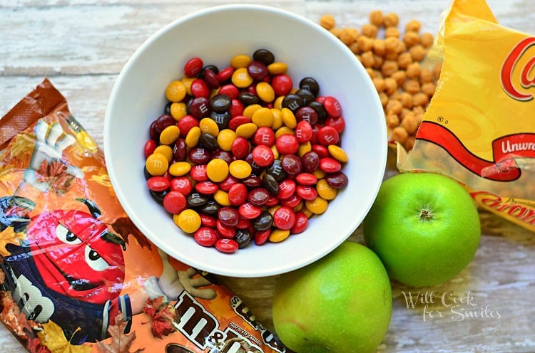 M&M bag and m&m's in a white bowl with 2 whole green apples to the bottom of it and a bag of caramel chips with some spilled out on the table 