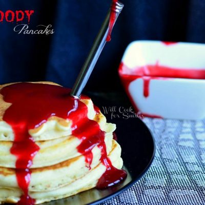 stack of pancakes with red fruit syrup as halloween decor blood to make bloody pancakes on black plate with fork stabbing pancakes and white square dish with more syrup