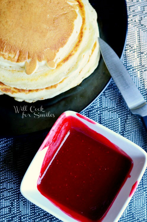 pancakes stacked up on a black plate with strawberry syrup in a small bowl 