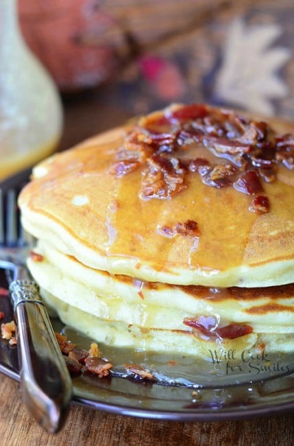 Maple Bacon Pancakes with bacon and syrup on top on a black plate with a fork to the left 