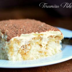 close up view 1 slice of tiramisu poke cake on decorative white dessert plate on wood table with fork in background to the right