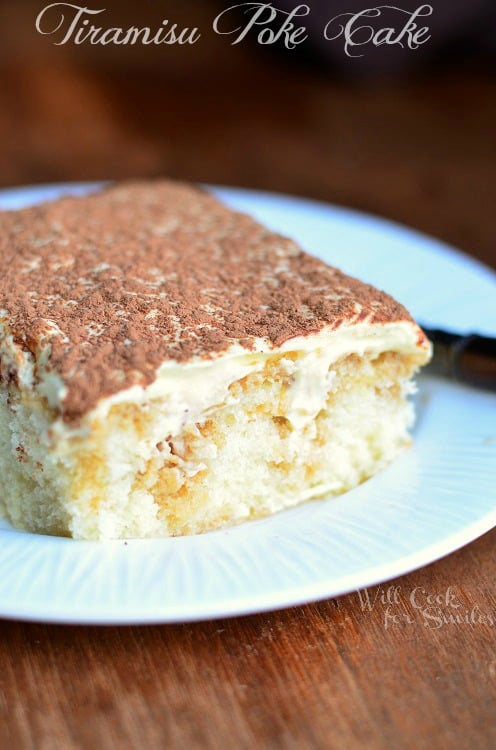 A piece of Tiramisu Poke Cake sits on a white, round dish. It's topped with mascarpone cream and dusted with cocoa powder.