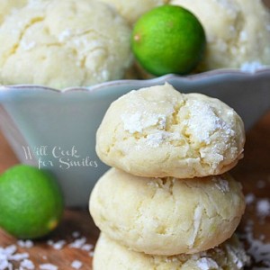 key lime cookies stacked up on a wood table with coconut shavings around it and a blue bowl in the background with cookies and limes
