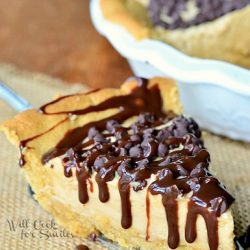 1 slice of peanut butter cookie cheesecake on cake cutter with rest of cake in background in white baking dish all on wood table