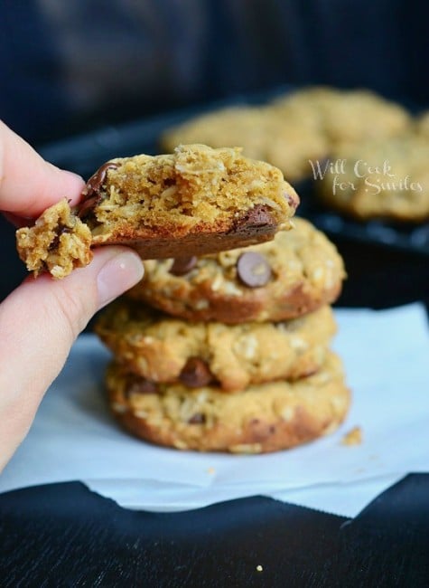 Cookies stacked up on wax paper on a black counter top and holding a cookie with a bite out of it 