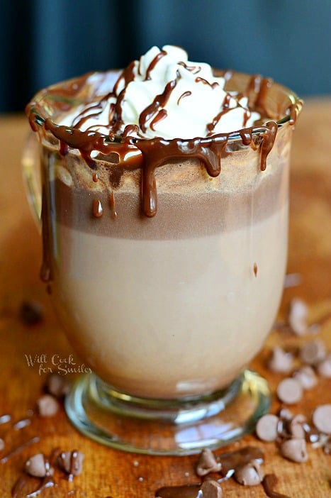 Hot Chocolate in a glass with whipped cream and chocolate sauce on a wood table with chocolate chips around it 