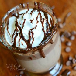 view from above of clear glass coffee mug filled with adult hot chocolate with whipped topping on a brown table with chocolate chips scattered around the bottom of the glass