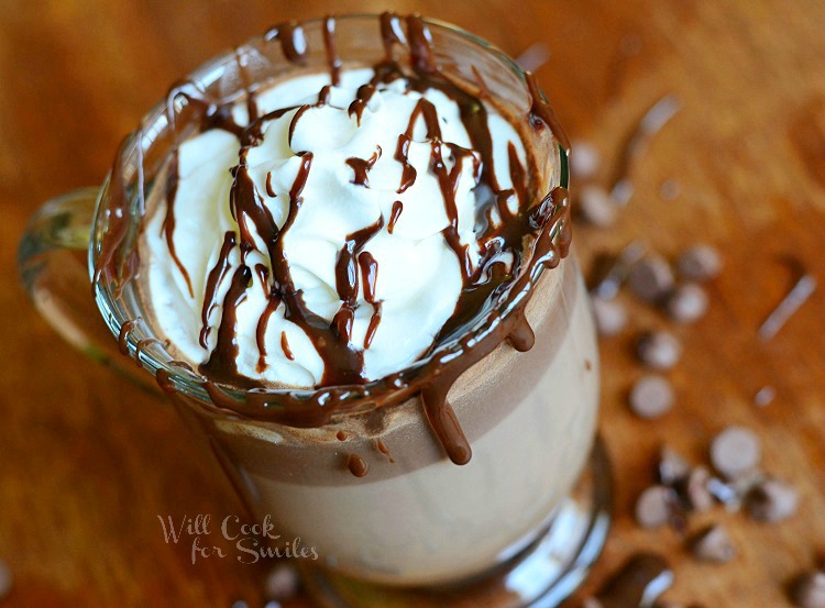 above photo of Hot Chocolate in a glass with whipped cream and chocolate sauce on a wood table with chocolate chips around it 