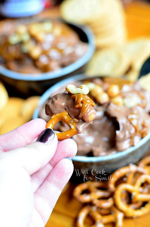 dipping a pretzel into Chocolate Fruit Dip in a blue bowl with pretzels to the right 