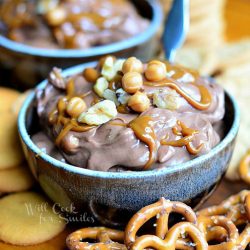 2 blue and black clay bowls filled with chocolate turtle dip with pretzles and cookies scattered around bowls and hershey's chocolate in glass bottles