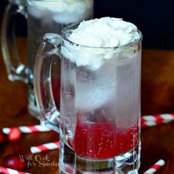 2 beer mugs filled with cranberry italian soda on wood cutting board with straws and cranberries scattered around mugs