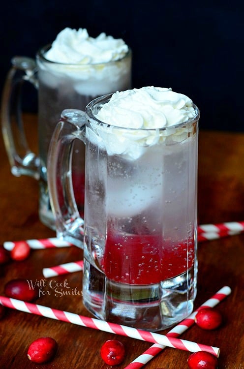 Cranberry Italian Soda in a glass mug with whipped cream in top and cranberries and red and white straws around the bottom on the wood table