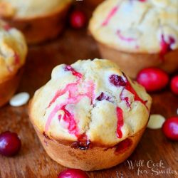 1 cranberry white chocolate chip muffin in foreground with additional muffins in background and cranberries and white chocolate chips scattered among muffins