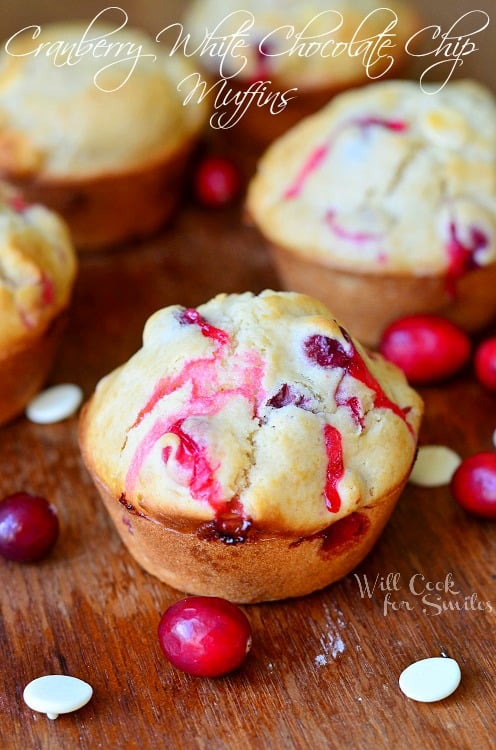 Cranberry White Chocolate Chip Muffins on a wood cutting board with cranberries and white chocolate chips scattered around them 