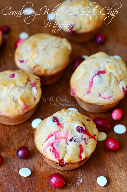 Cranberry White Chocolate Chip Muffins on a wood cutting board with cranberries and white chocolate chips scattered around them 