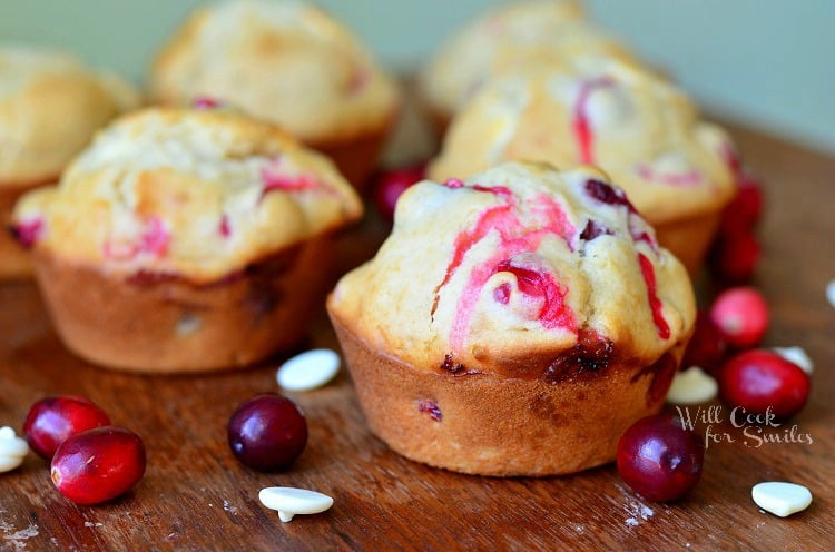 horizonal photo of Cranberry White Chocolate Chip Muffins on a wood cutting board with cranberries and white chocolate chips scattered around them 